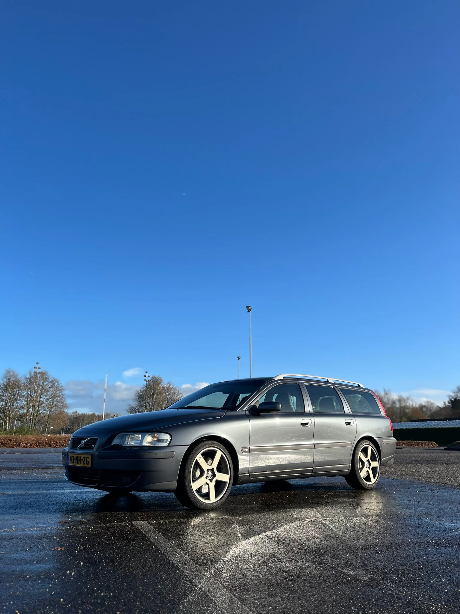 Grijze Volvo V70R op een nat parkeerterrein met een grote blauwe lucht erboven