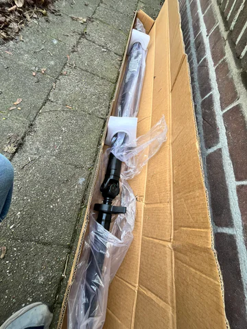 Nieuwe NTY-aandrijfas in een lange kartonnen doos naast een bakstenen muur